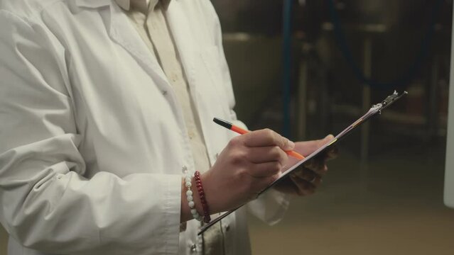 Tilt up of Biracial female brewery supervisor taking notes in checklist on clipboard while standing by control board and pressing buttons, checking work of machine equipment at factory