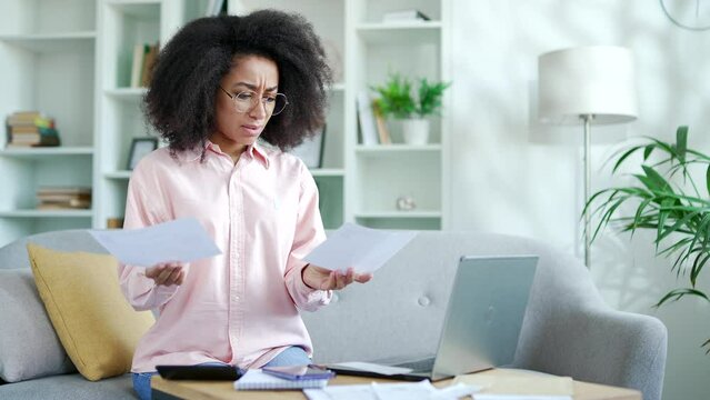 Confused young african american female having difficulty with paper work doing household budget sitting on sofa in living room at home office. Puzzled shocked black woman looks at large utility bills