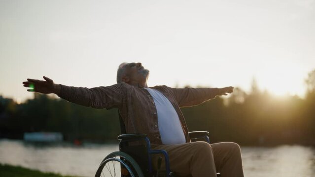 Senior man in wheelchair enjoys freedom. Male with special need spreading arms on sides smiling breathing fresh air walking in the city park. Living with physical disability in old age concept.