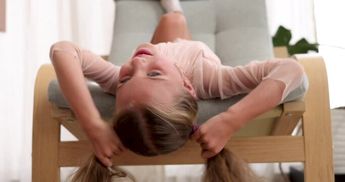 Smiling preschooler girl with ponytails hangs head down in armchair in premise of apartment. Entertainment from boredom at home in childhood slow motion