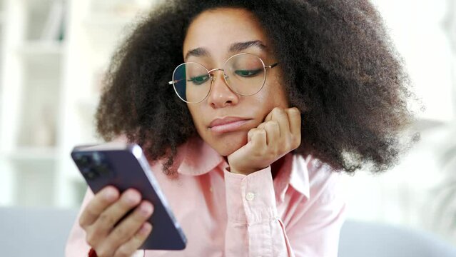 Depressed sleepy young african american female bored using smartphone sitting on sofa in living room at home. Sad lazy lonely black woman student yawns, has a bad mood, browses mobile phone. Close up