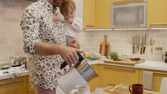 Tilt up of bearded father in stylish shirt standing in kitchen and holding cute toddler girl sucking her thumb he pouring water from electric kettle into baby bottle