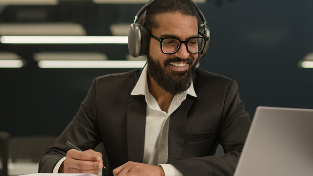 Happy smiling satisfied Arabian man business manager entrepreneur employer Indian positive smile businessman writing notes in notebook wear headphones online lesson class video call studying in office