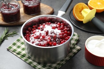 Making cranberry sauce. Fresh cranberries with sugar in saucepan and ingredients on gray table, closeup