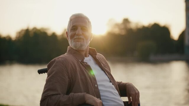 Senior man in wheelchair enjoys freedom. Male with special need look at the camera smiling breathing fresh air walking in city park near pond. Living with physical disability in old age concept.