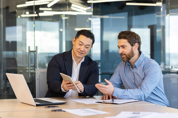 Two professional businessmen engaged in a discussion over digital tablet and laptop in a corporate...