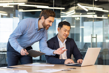 Two professional male colleagues collaborate at a work desk in a bright modern office, discussing...