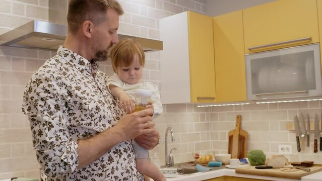 Tilt up of caring bearded father in stylish shirt standing in kitchen and holding toddler girl, giving her baby bottle with diluted juice