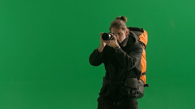 A male traveler takes pictures with a camera while hiking. A tourist with a backpack on his back stands in a studio on a green screen. Concept of travel, active rest, hiking.