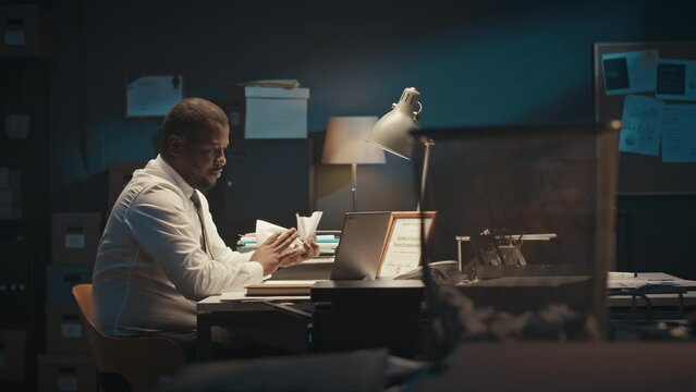 Medium shot of stressed Black male executive sitting at desk at night time, crumpling and throwing sheets of paper into bin, working overtime alone in dark office