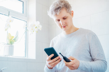 Young man puts the settings of his electronic toothbrush with mobile phone app. Wireless connecting sonic toothbrush with smart phone app. Modern home health care technology concept. Selective focus