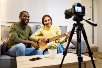 A dark-skinned couple is sitting on the couch at home playing a flamenco guitar and singing while...