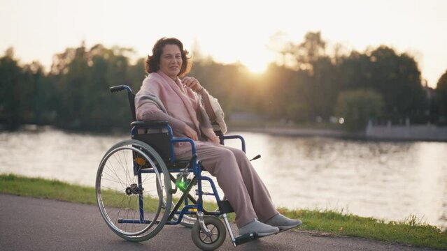 Woman in wheelchair walking in city. Senior mature smiling woman breathing air outdoor thinking looking around enjoying pastime at sunset. Live with physical disability, special needs people concept.
