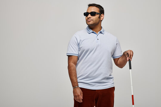 attractive indian blind man in blue tee shirt with glasses and walking stick posing on gray backdrop