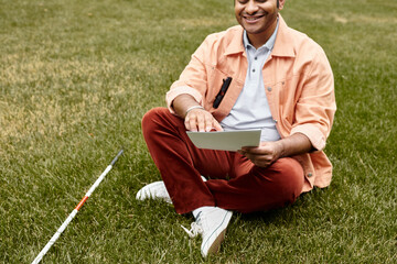 joyful indian man with blindness in orange jacket sitting on grass and reading braille code
