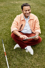 cheerful indian man with blindness in orange jacket sitting on grass and reading braille code