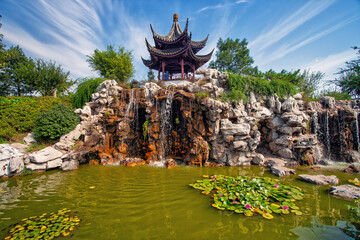 an old chinese pavilion with rocky waterfalls and a pond with water lilies