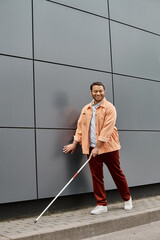 joyous indian blind man in orange jacket with helping stick walking with gray wall on backdrop