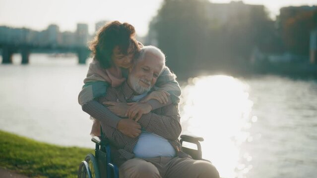 Wife walking with husband in wheelchair. Senior family couple spending time in city park. Woman hugging man showing love care, they enjoying life. Living with physical disability, care concept.