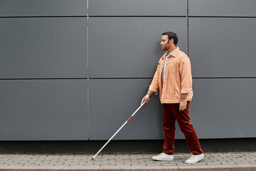 attractive indian blind man in orange jacket with helping stick walking with gray wall on backdrop