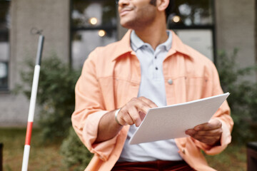 cropped view of indian man with visual impairment in vivid jacket on bench reading braille code