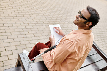 cheerful indian blind man in orange jacket sitting on bench and reading braille code while outside