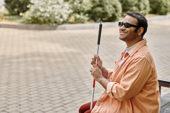 merry indian blind man in orange jacket sitting outside on bench with walking stick and glasses