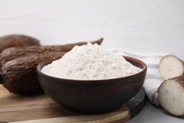 Bowl with cassava flour and roots on white table, closeup
