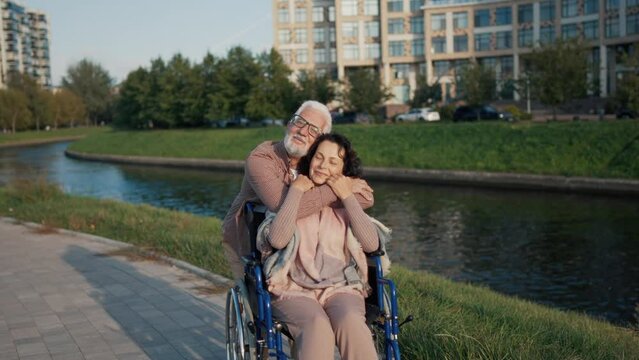 Husband hugging his wife in wheelchair. Senior happy family spending time in city park near pond taking photos on smartphone. Couple showing old love care. Living with physical disability concept.