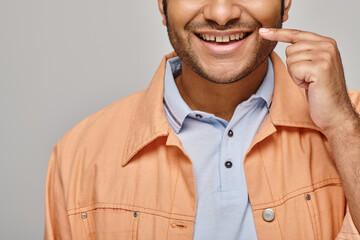cropped view of joyful indian man in orange jacket posing with finger near face on gray backdrop