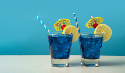 Two blue cocktails in a glass filled with ice garnished with lemon slices, cherries, yellow...