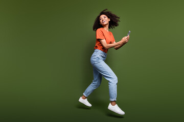 Fototapeta na wymiar Full length photo of cheerful dreamy lady dressed orange t-shirt jumping typing modern gadget empty space isolated khaki color background