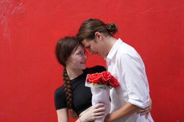 couple young man and woman hugging and holding the red bouquet rose flowers at red wall background....