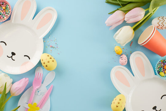 Easter delight fiesta: kids' exuberance with egg-citement. Top view photo of bunny shaped plates, eggs, paper cups, cutlery, tulips, sprinkles on pastel blue background with space for festive text