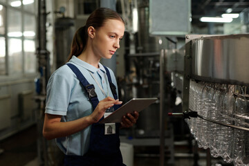 Young quality control staff with tablet looking at empty plastic bottles in factory conveyor and...