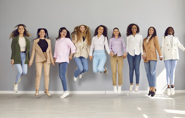 Naklejka premium Group of funny business multiethnic women looking at camera, holding hands smiling and jumping on a grey wall background. Company employees happy to make a good deal. Team work concept.