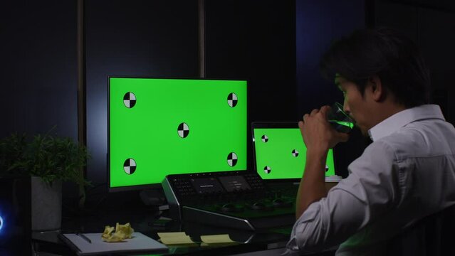 Young Asian man with longer hair using two computers screens with green screen on