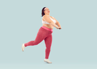 Full length shot of funny chubby overweight woman doing sports. Happy plus size, fat woman wearing sportswear doing physical exercises isolated on light blue studio background