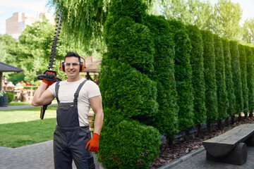 Muscular male gardener in uniform, safety glasses and gloves holding electric hedge trimmer in hands. Handsome caucasian man smiling and looking at camera while standing on back yard. - Powered by Adobe