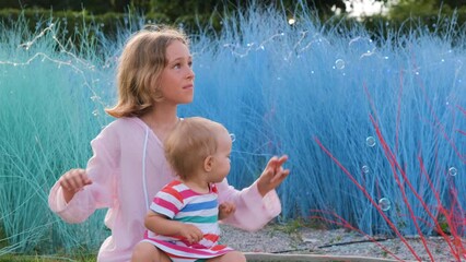 Little girl with cute baby sister catching soap bubbles by the colorful grass in the evening, slow motion. Happy childhood concept