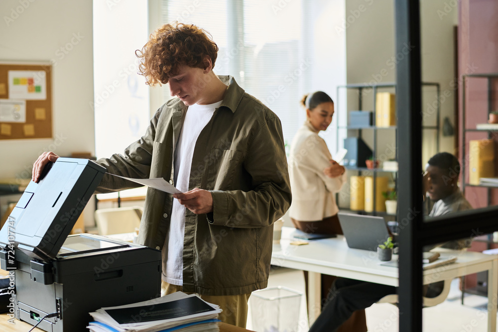Poster young office manager in casualwear looking at financial paper while standing in front of xerox machi