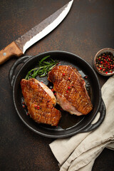 Two roasted duck breast fillets with crispy skin, with pepper and rosemary, top view in black cast iron pan with knife, dark brown concrete rustic background.