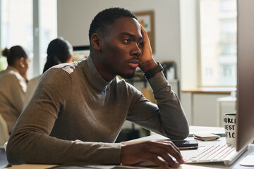 Young bored African American businessman sitting by workplace in front of computer and looking at...