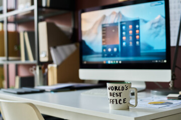 White porcelain mug with note world best office standing on desk of broker or designer against computer monitor with blue screen