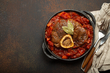 Traditional Italian dish Ossobuco all Milanese made with cut veal shank meat with vegetable tomato sauce served in black casserole pan top view on rustic brown background, copy space.
