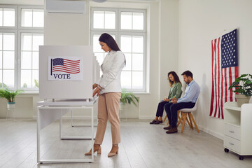 Side view portrait of young american voter woman standing at vote center with USA flags in voting...
