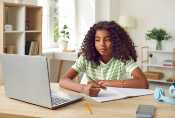 Smiling African American Girl studying with laptop computer. Teenage girl sitting at her desk and writing in notebook. Student doing her homework or learning online. Remote education or homeschooling
