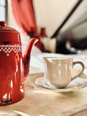 A white porcelain cup and a red coffee pot in blurry focus stand on a wooden tray on a white bed lit by bright morning light.
