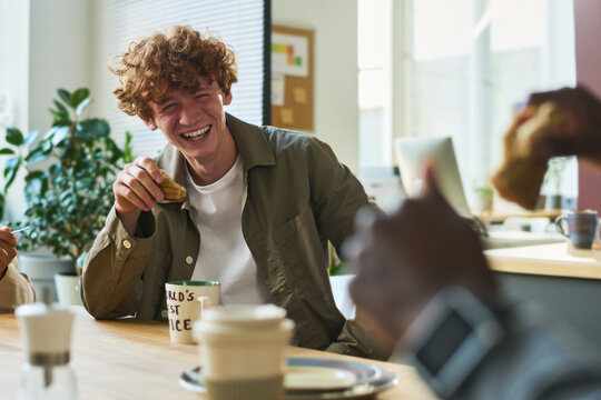 Young laughing male office worker with cookie or some other snack in hand sitting in front of colleague and enjoying lunch with him