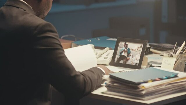 Back view chest up of African American workaholic businessman studying paperwork and looking at family portrait in picture frame on desk while overworking in office alone late at night